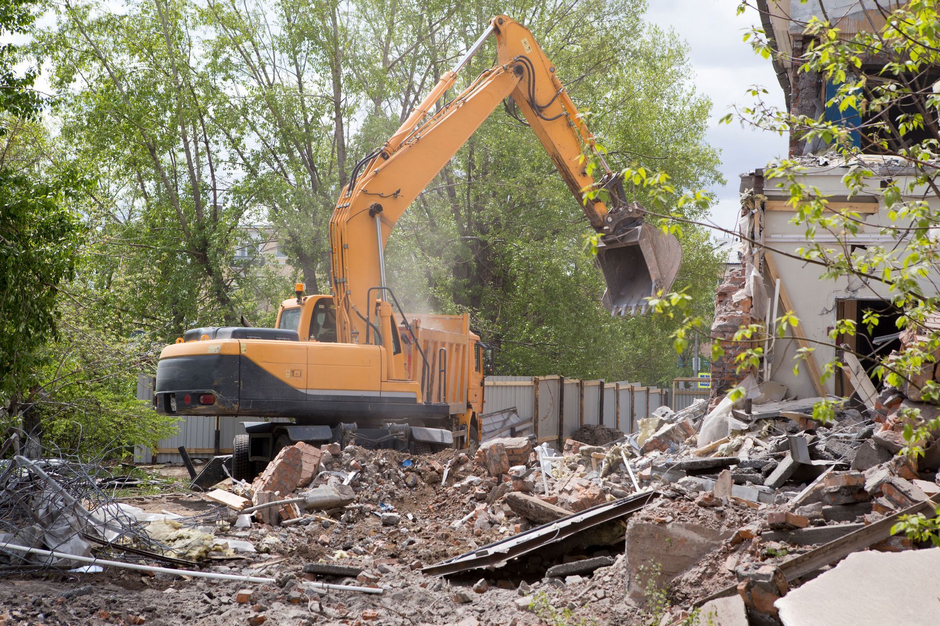 Yellow excavator demolishing rubble beside a building in a debris-filled lot