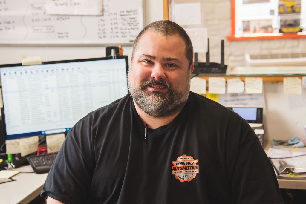 Man sitting at an office desk, smiling in front of computer monitors and paperwork.
