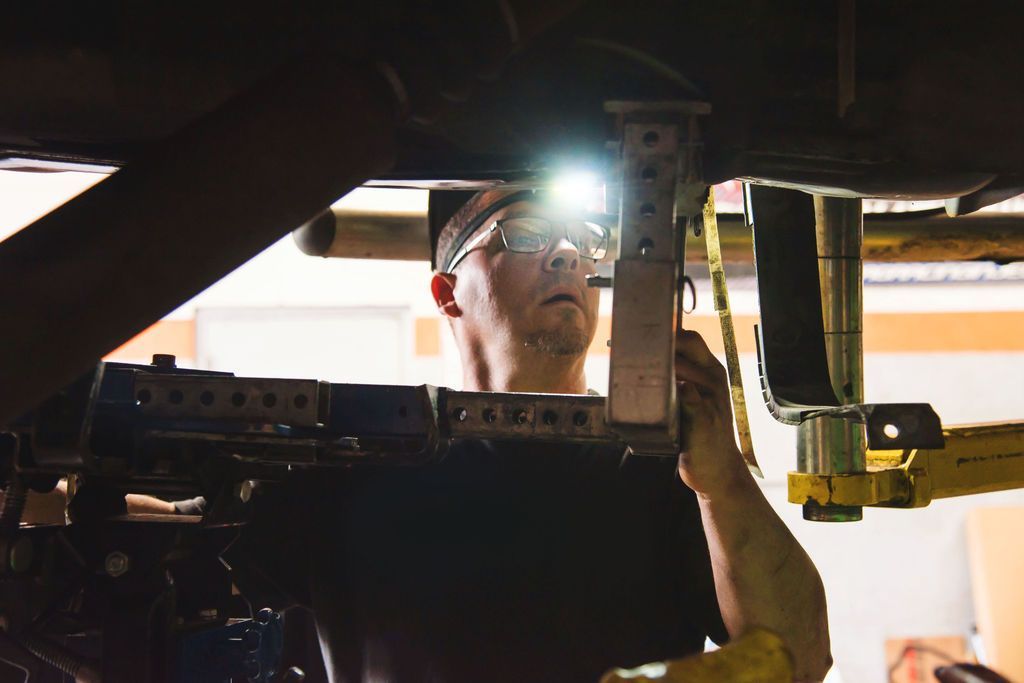 Worker inspecting machinery under a vehicle, wearing a headlamp and safety glasses.