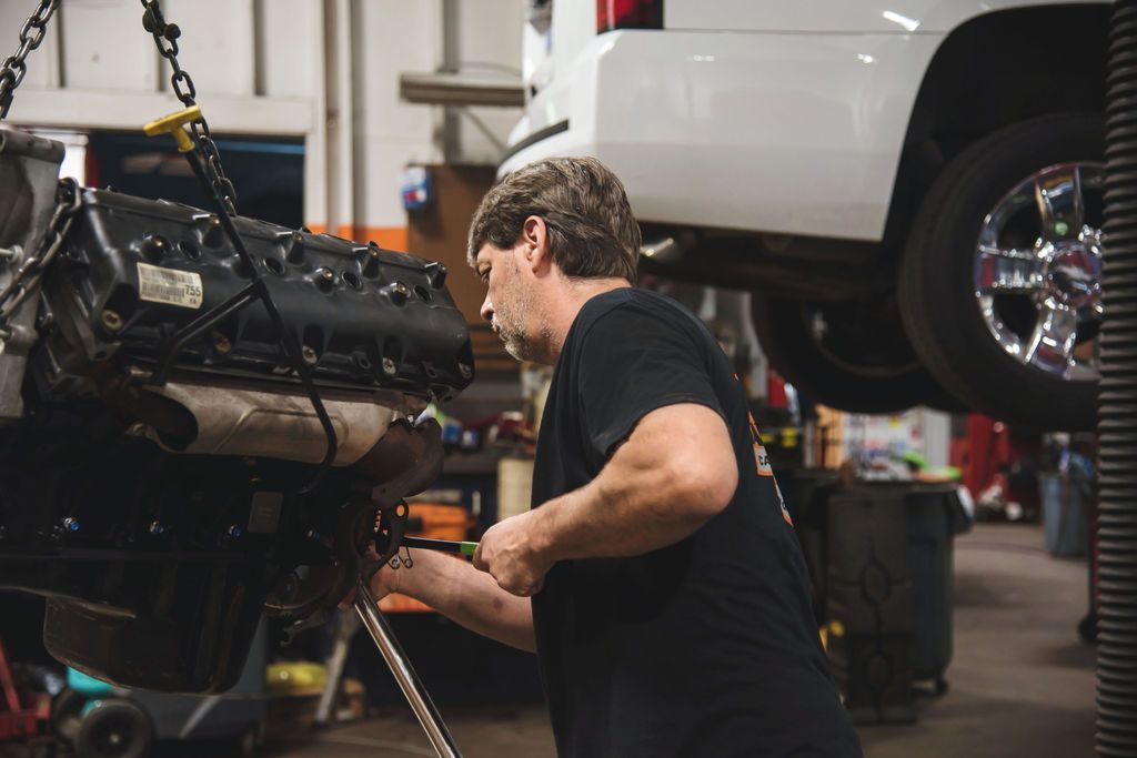Mechanic working under a lifted white SUV in an auto shop, focused on engine repairs