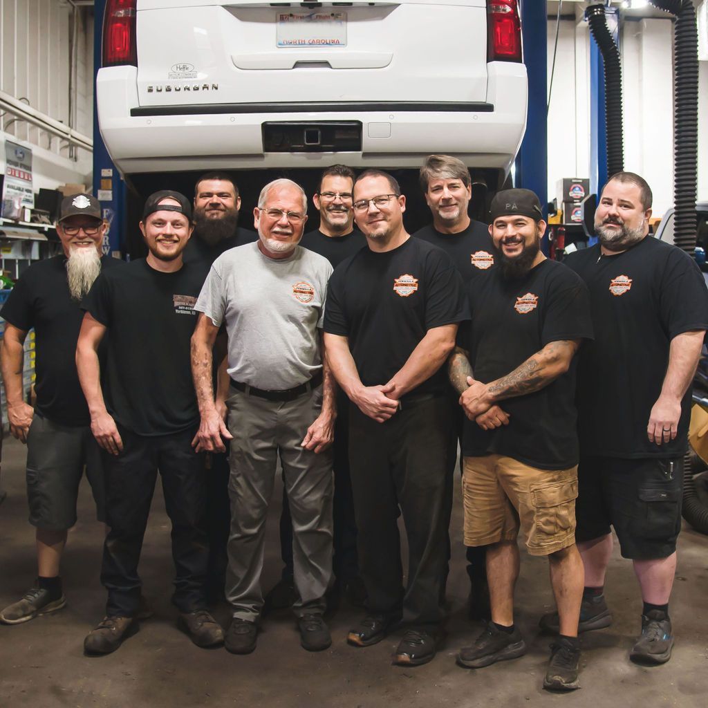 Group of auto shop workers standing in a garage under a raised white van, smiling at the camera.