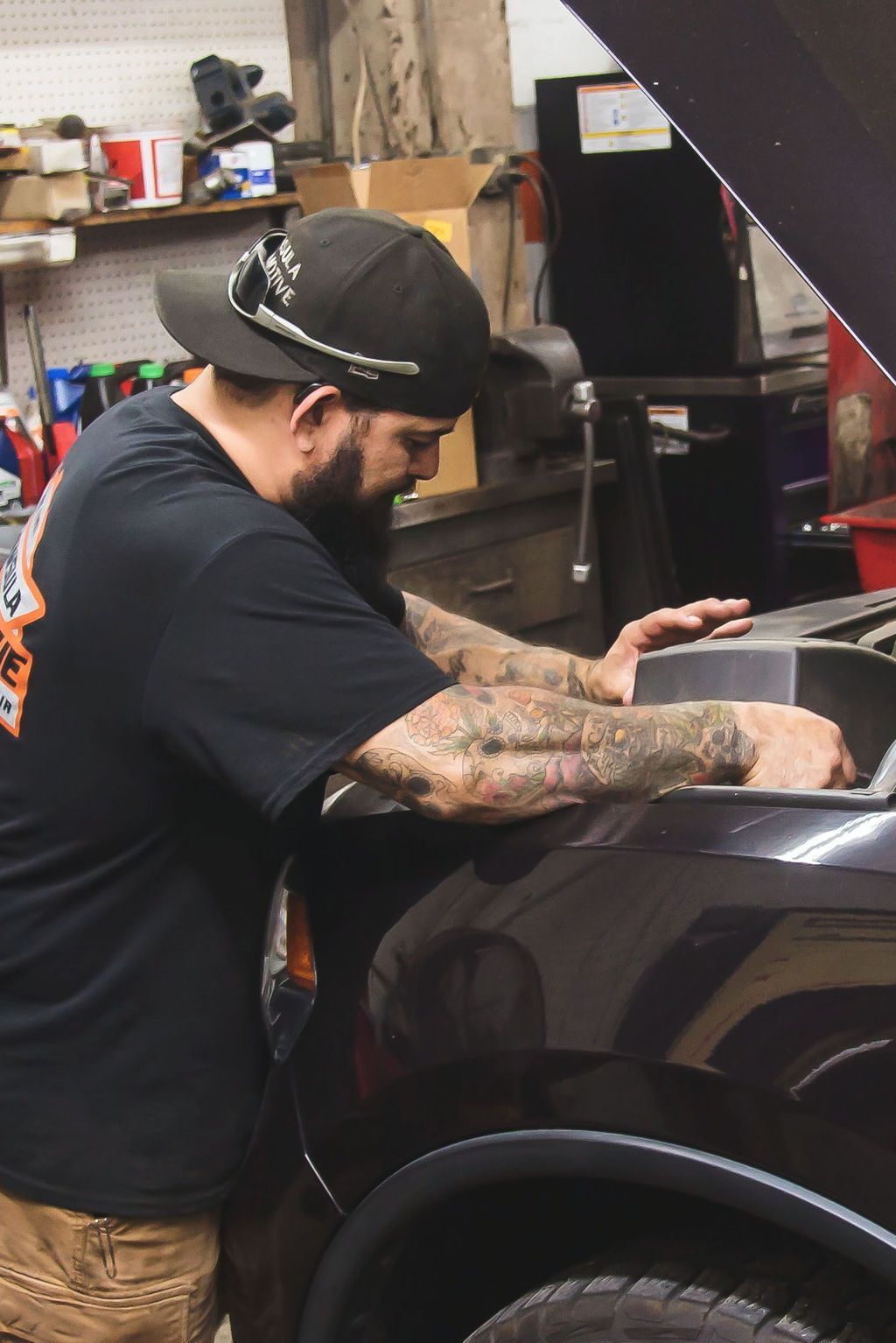 Man leaning over a black car in a garage, working on the front fender with tattooed arms visible