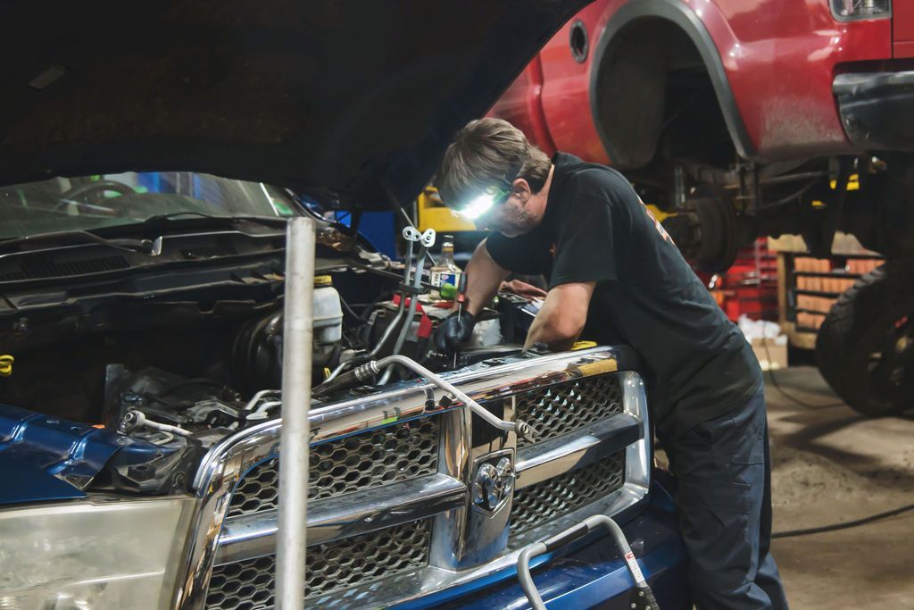 Mechanic inspecting an engine in a garage, with a blue car and a red truck on a lift behind him