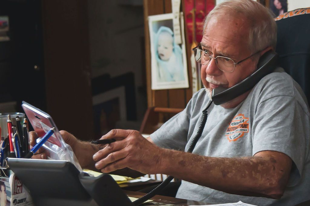 Man seated at a desk, speaking on a phone while using a tablet and stylus in a dim office.