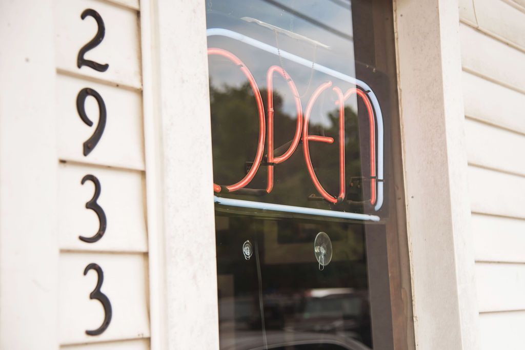 Neon “open” sign in a storefront window beside the number 2933 on white siding