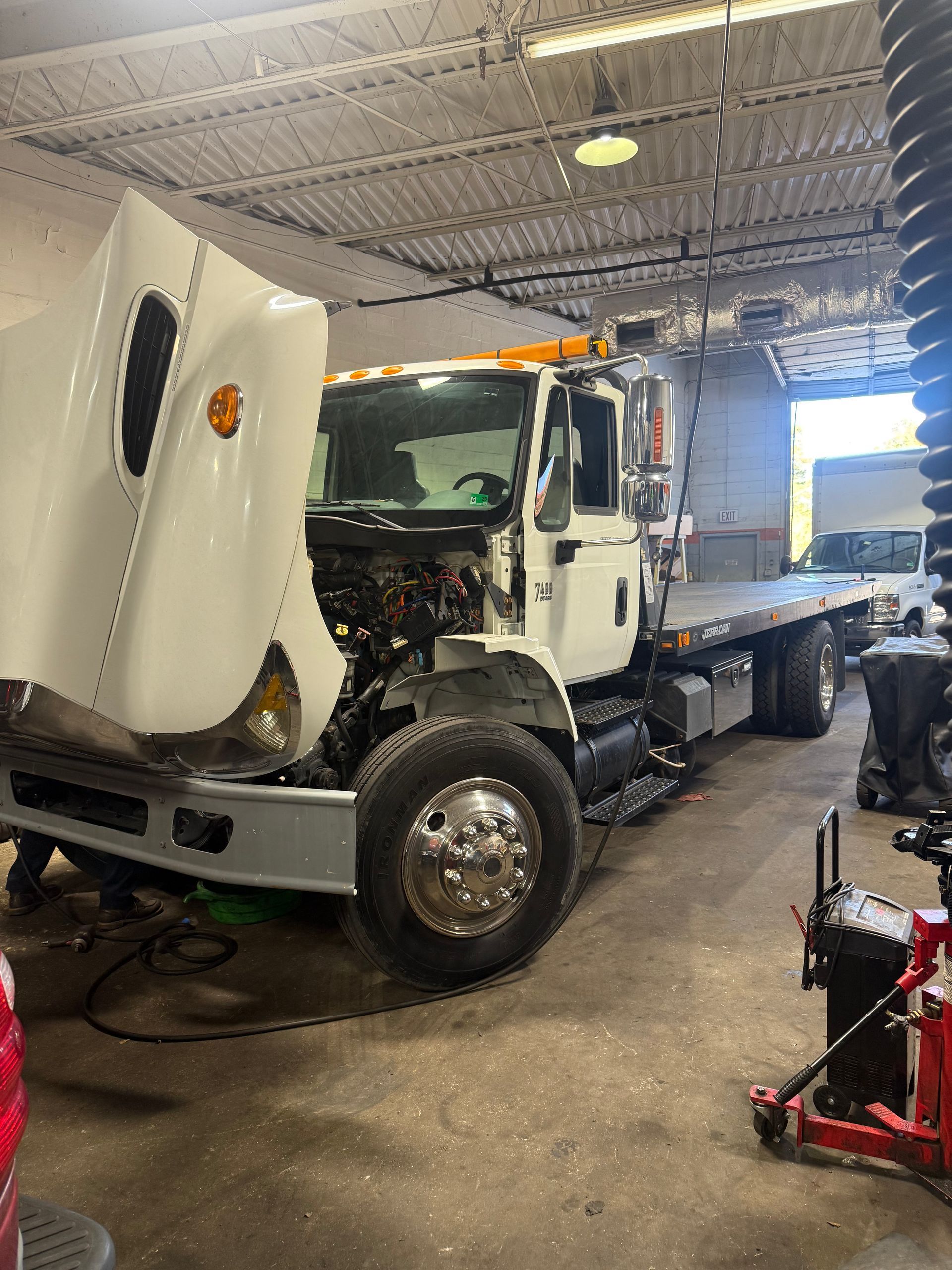 White tow truck with raised hood in a garage bay, front view, surrounded by tools and equipment