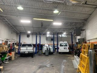 Auto repair shop with vehicles on lifts, a white van, and yellow safety barriers inside a garage
