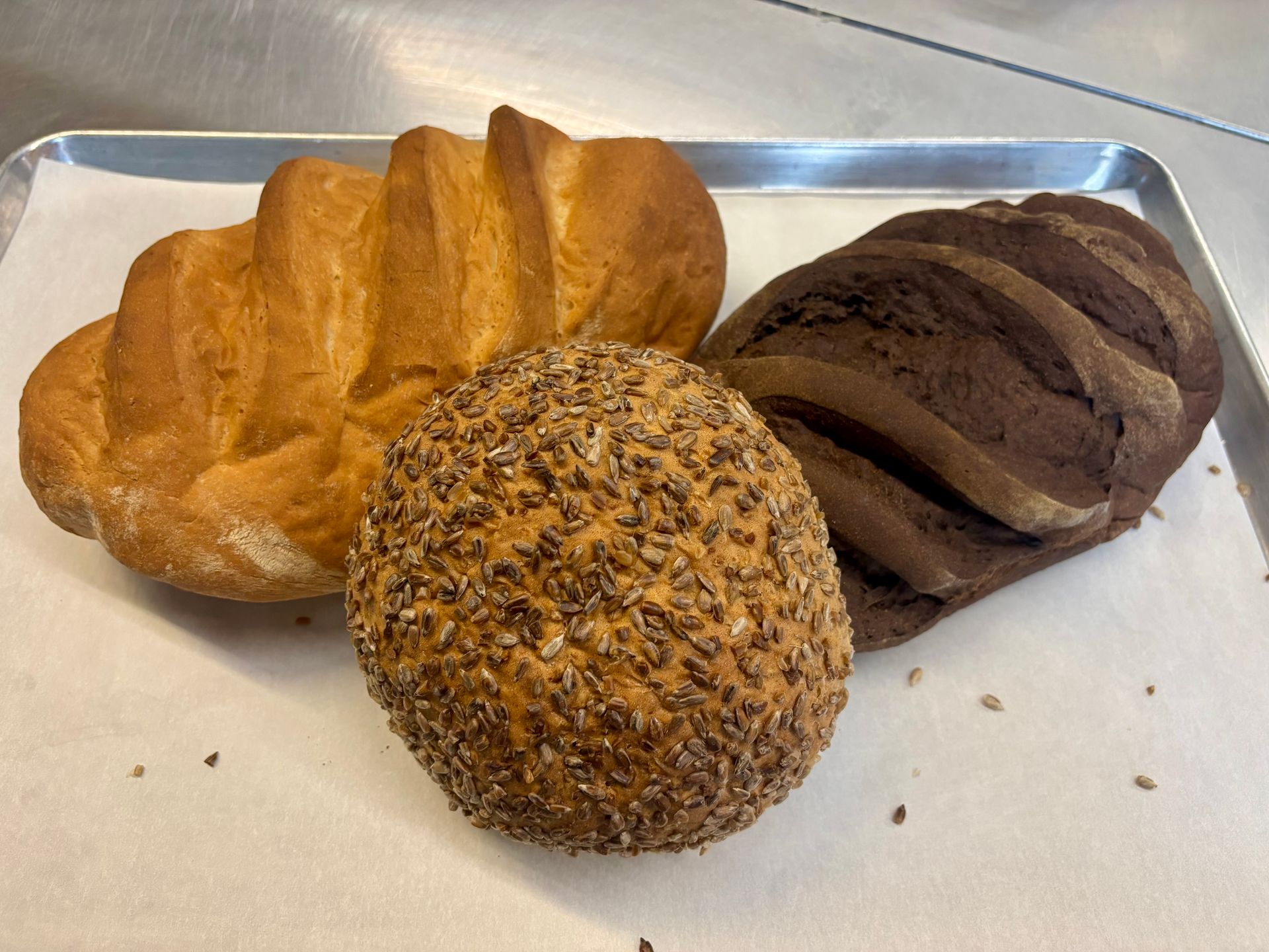 Three assorted bread rolls on a metal tray, including sesame-coated and dark rye bread.