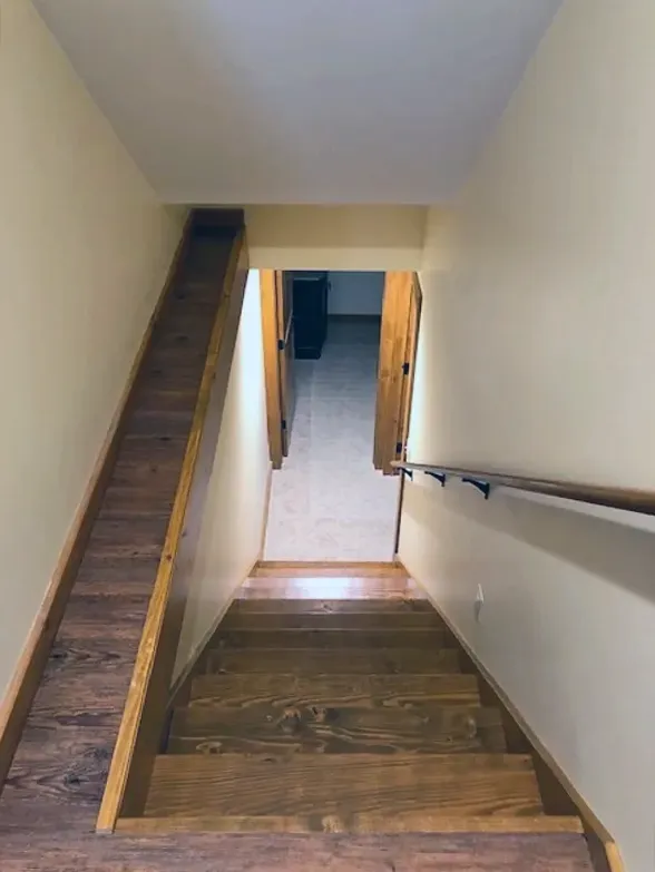 Stairwell with wooden steps and beige walls, leading down to a doorway and handrail.