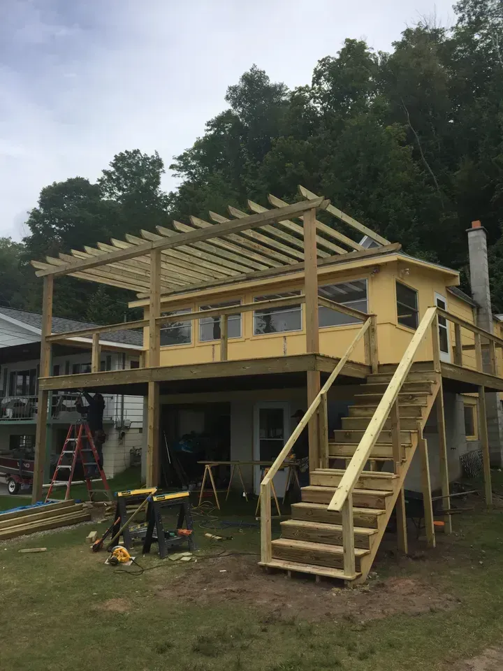 Partially built yellow house with a wooden deck, stairs, and roof framing in a yard