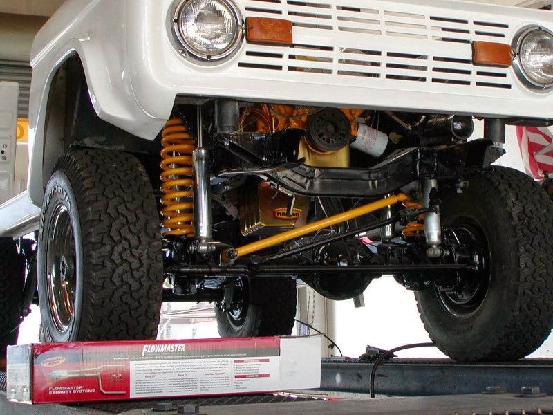 White vintage Ford Bronco, lifted with off-road suspension. Shows engine bay and front tires, with a box of parts in front.
