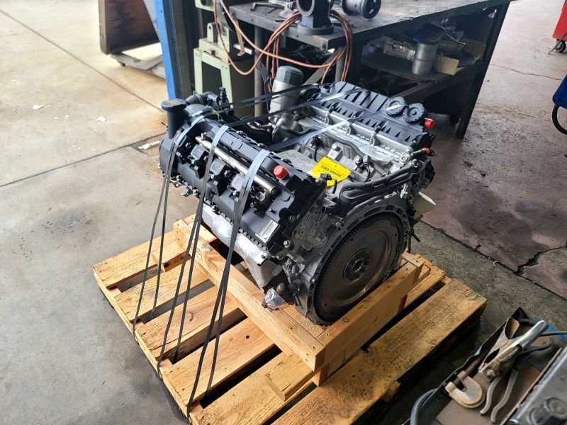 Engine on a wooden pallet in a repair shop. Black engine block with various attached components, secured with tie-downs.