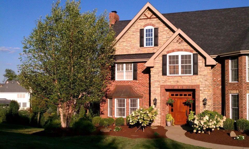Brick house with a brown door, black shutters, and well-manicured landscaping under a blue sky.