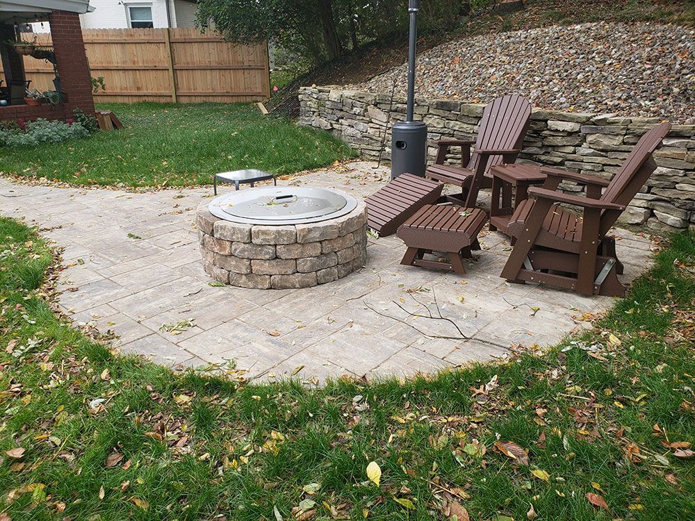 Stone fire pit and seating area with chairs on a stone patio in a backyard setting.