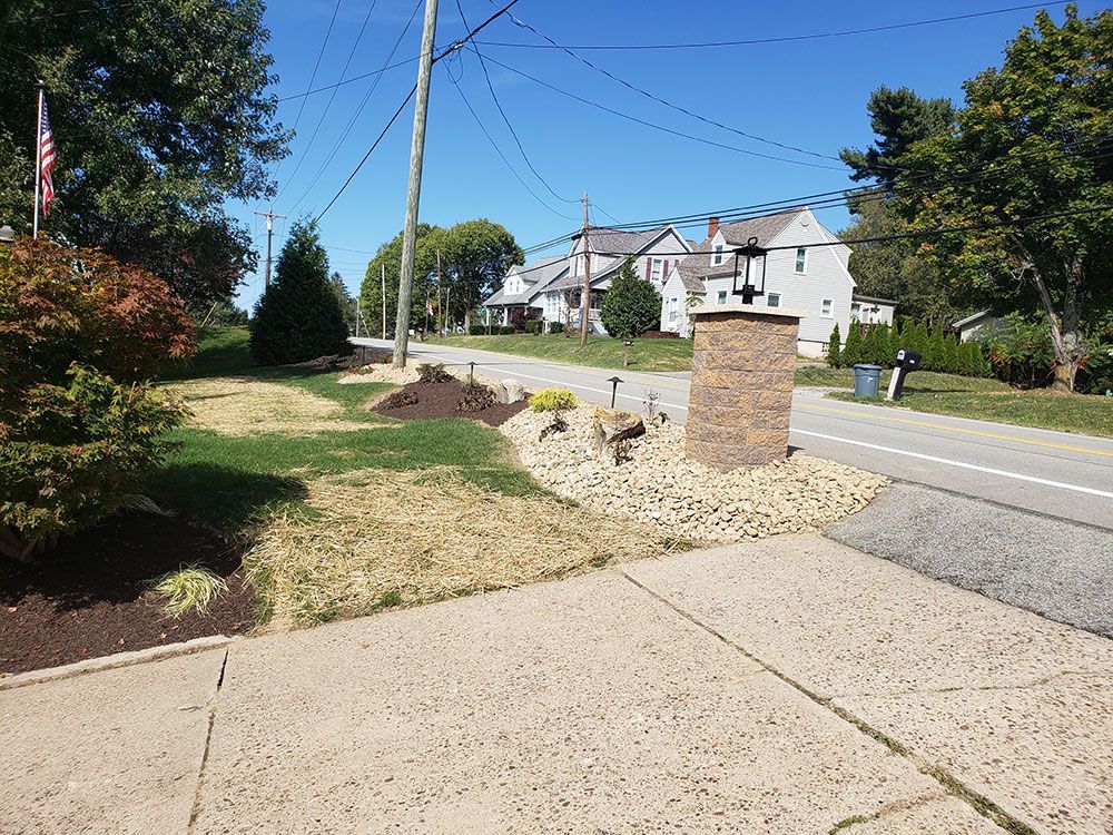 Residential street scene: Brown stone pillar in front yard, dry grass, houses in the distance, blue sky.