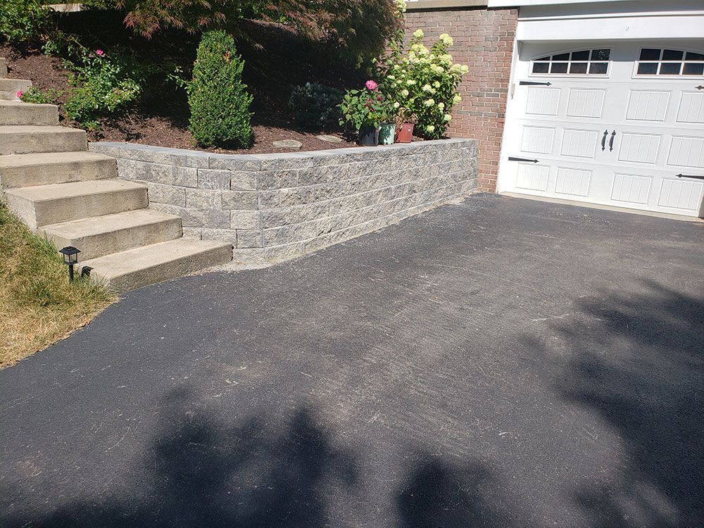 Stone retaining wall with steps and driveway. Plants are in the garden bed. Garage is on the right.