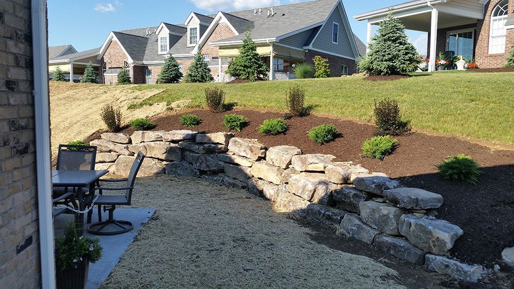 Stone retaining wall with a landscaped yard, patio furniture, and houses in the background.