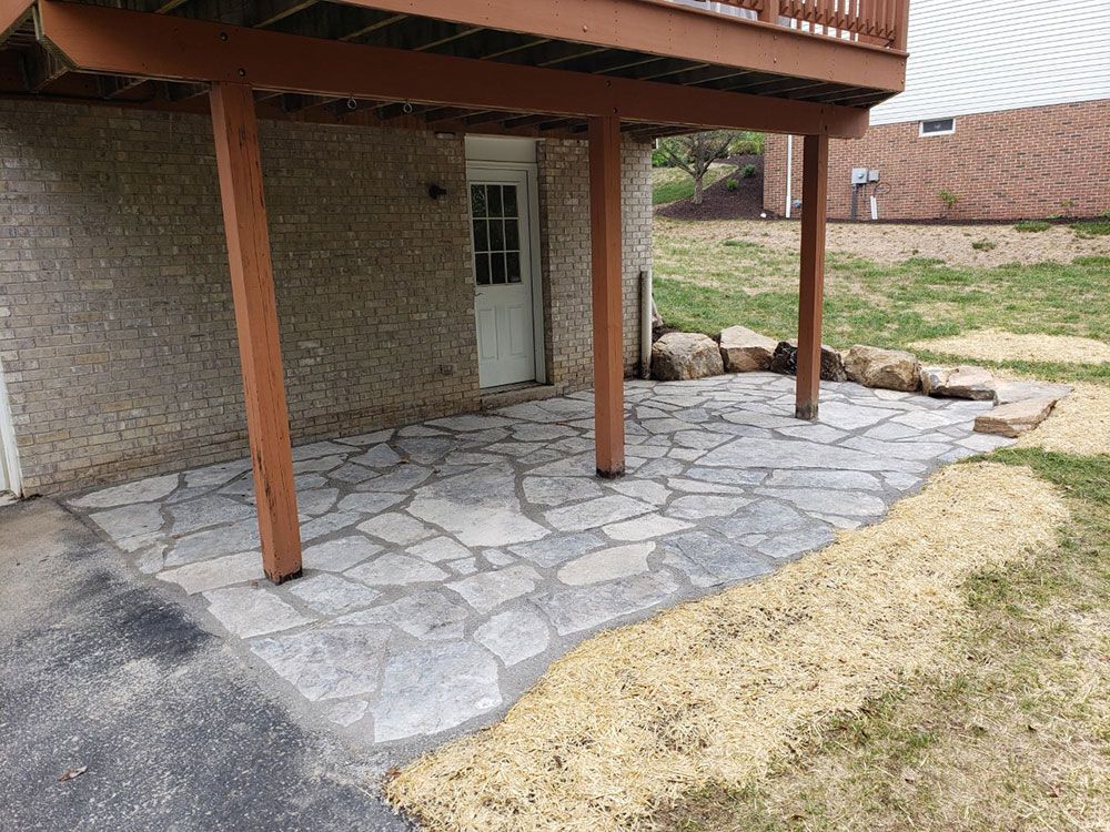 Stone patio under a wooden deck supported by brown posts. A white door is visible in the brick wall.