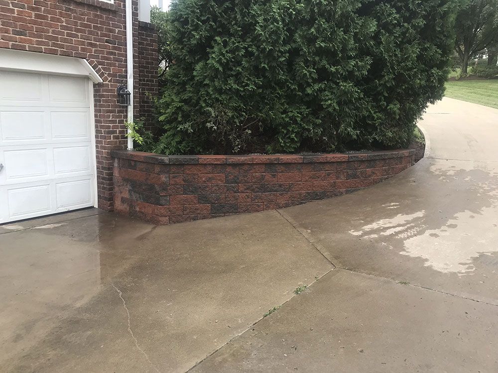 Brick retaining wall next to a garage and driveway with a large evergreen bush.