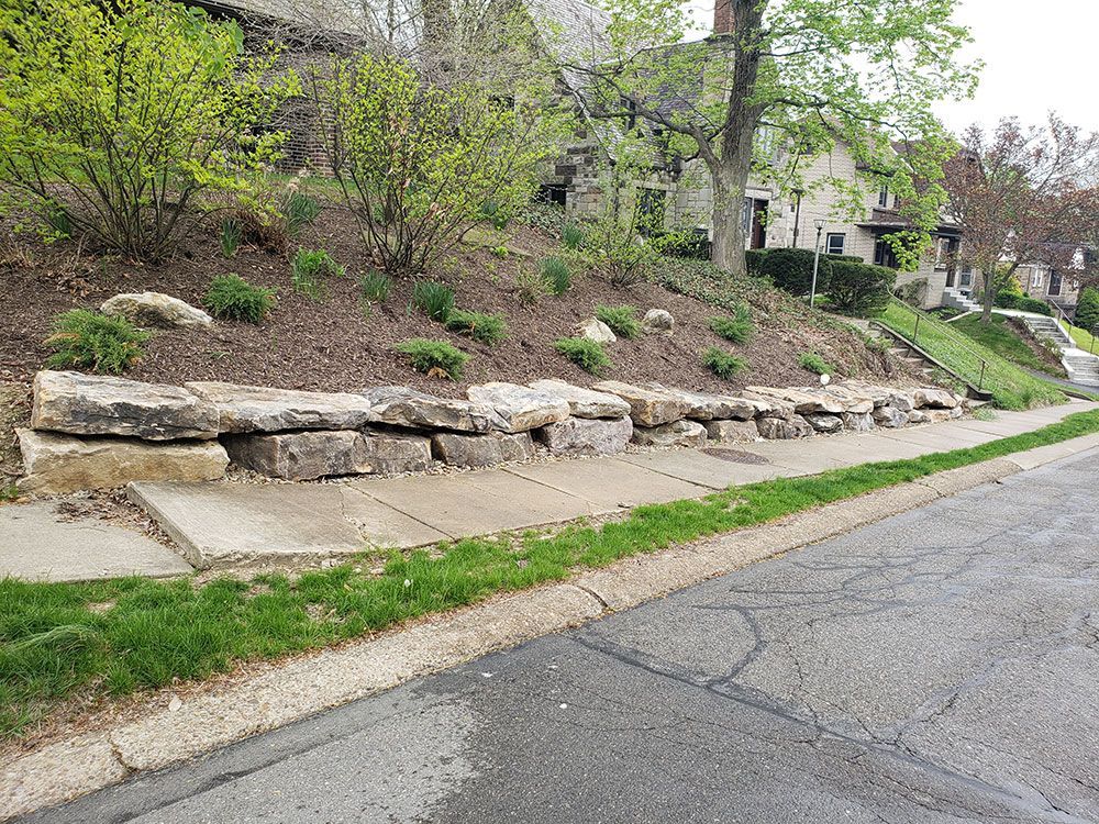 Stone retaining wall next to a sidewalk on a grassy hillside. Trees and bushes in background.