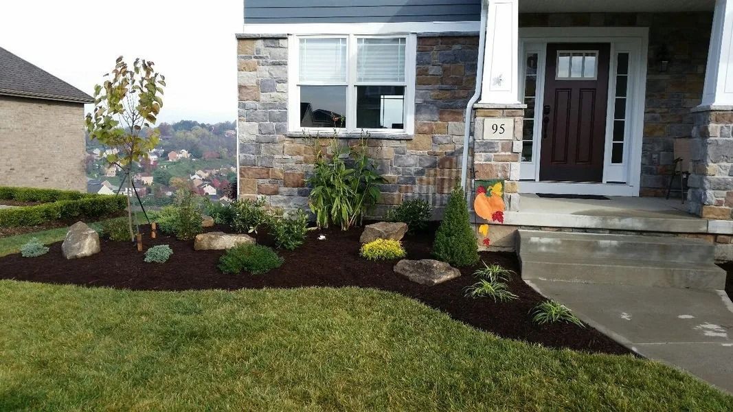Landscaped yard with mulch, plants, and large stones in front of a stone-faced house.