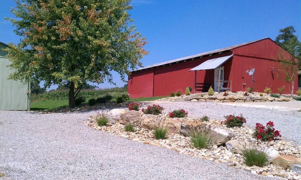 Red barn with white roof and a rock garden on a gravel drive with green grass and trees.