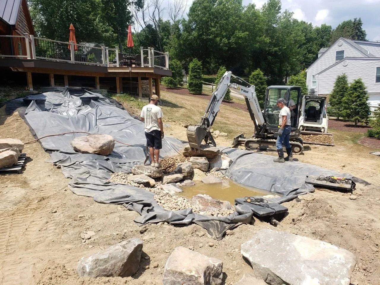 Men using heavy machinery to build a pond in a yard with a deck and house in the background.