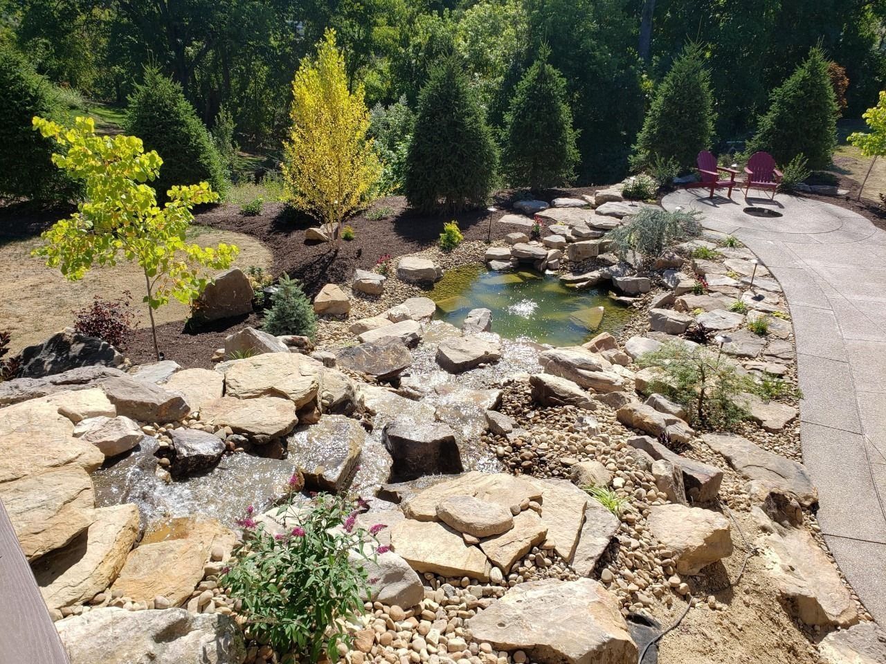 Landscaped yard with a pond, waterfall, and pathway surrounded by rocks and trees.