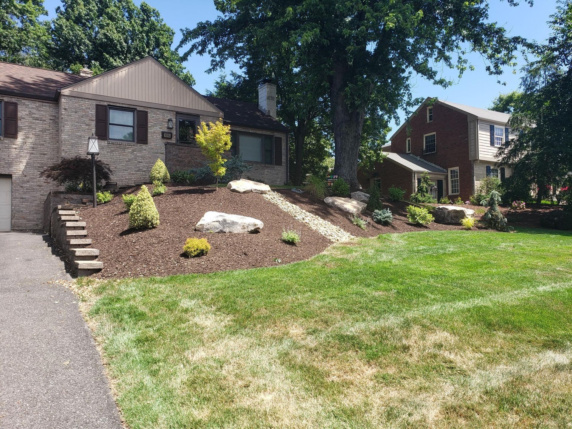 Landscaped yard with stone and mulch in front of two-story brick homes and green lawn.