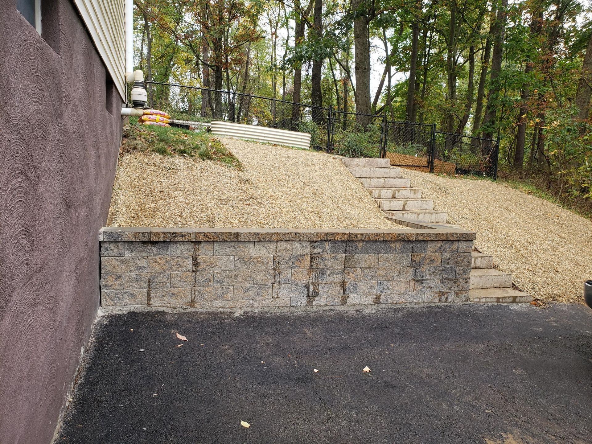Stone retaining wall with steps leading up a gravel hill to a wooded area. Asphalt in foreground.