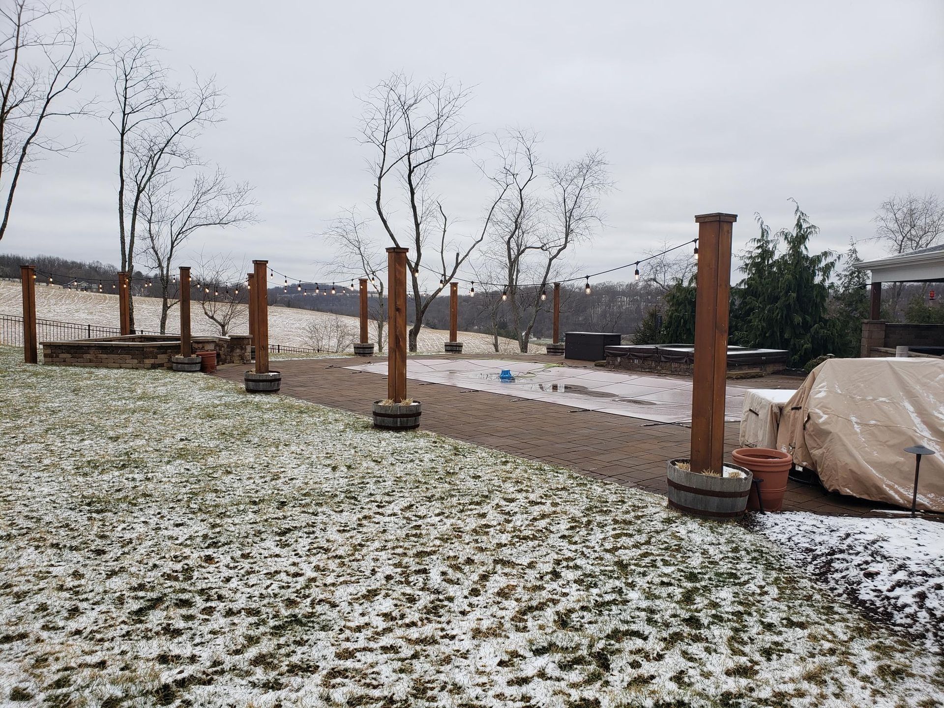 Snow-covered yard with wooden posts and path leading to a covered area, trees in the background on a cloudy day.