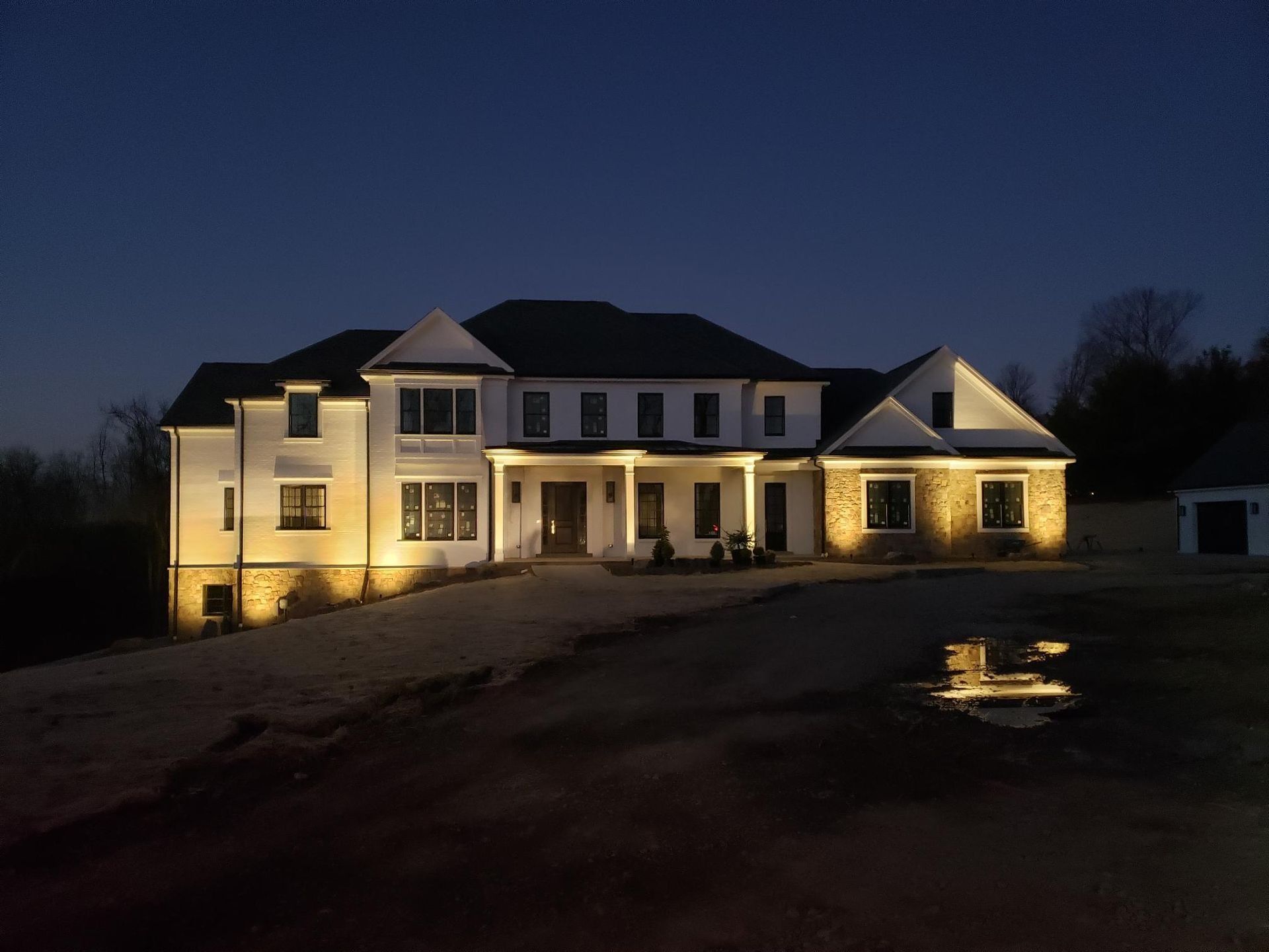 Large, two-story house illuminated at night with warm lighting. Features stone and white siding with a long driveway.
