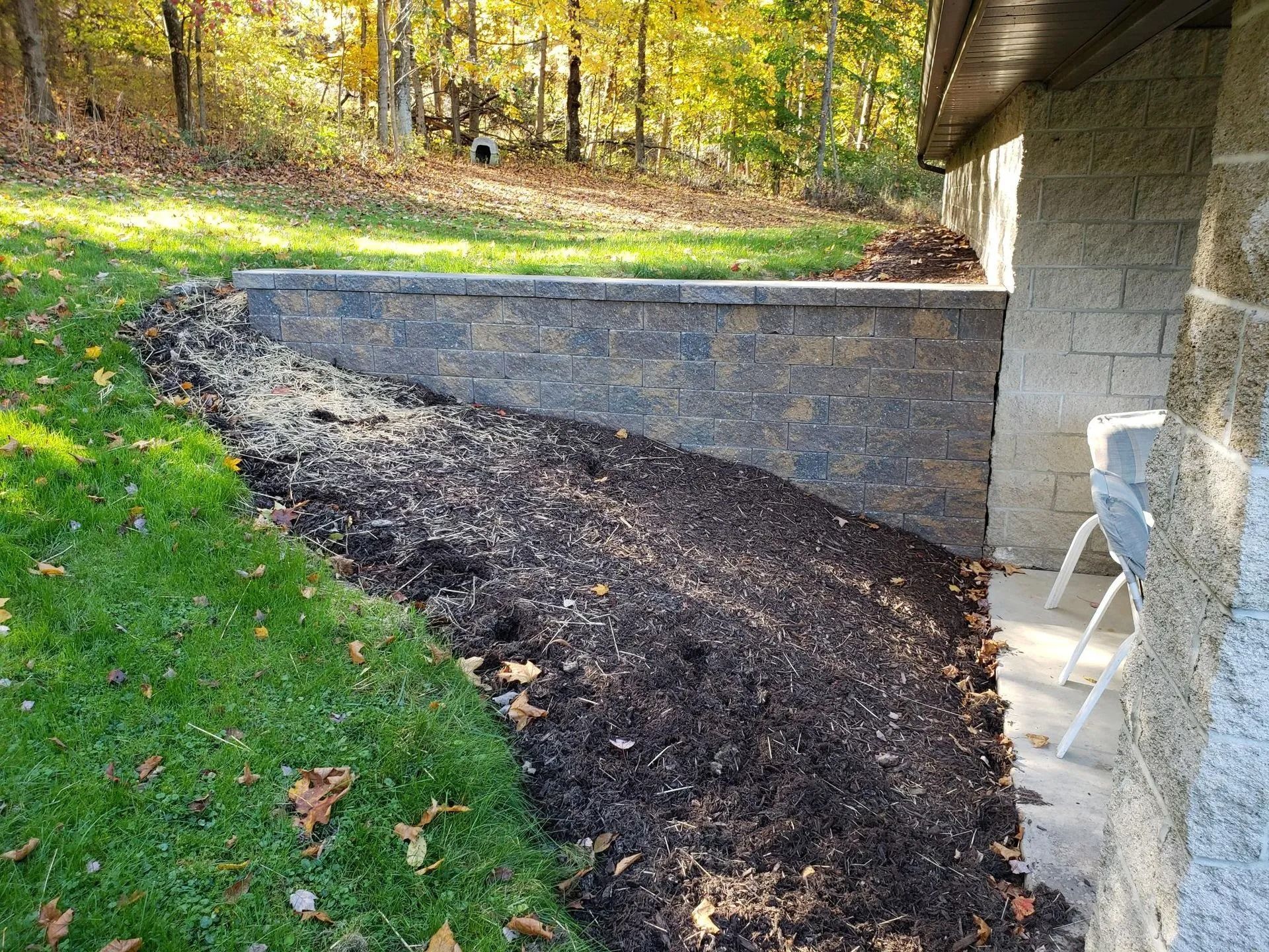 Retaining wall with mulch, beside building and grassy slope; autumn leaves present.