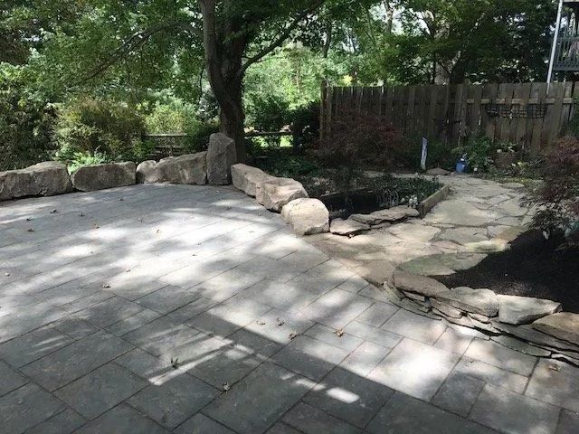 Stone patio and walkway lead to a pond in a backyard with trees and a wooden fence.