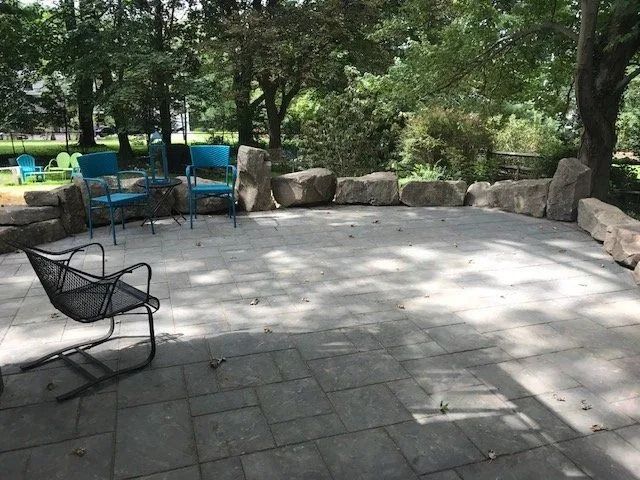 Stone patio with blue chairs and a black chair, surrounded by large rocks and trees.