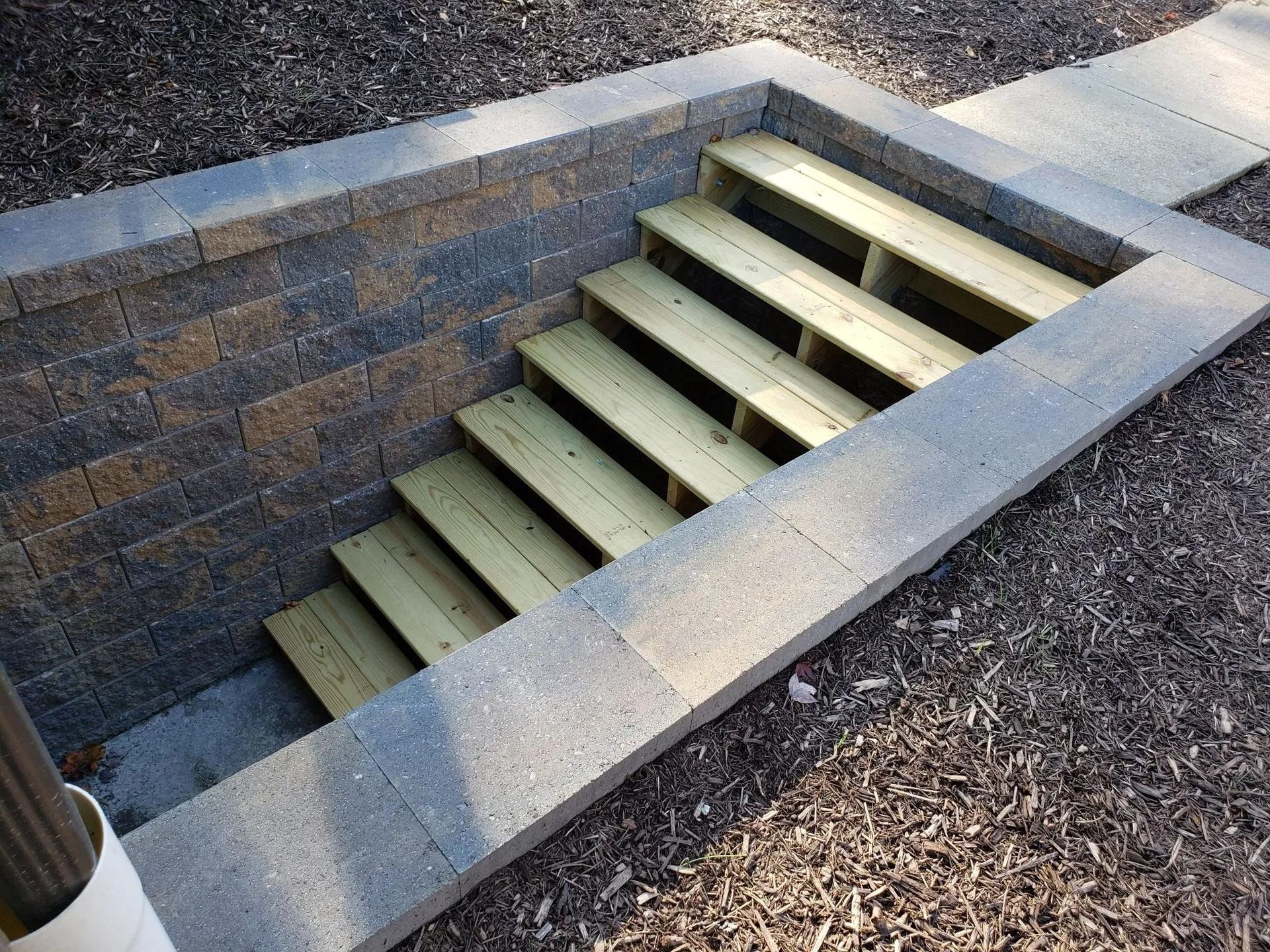 Stone-walled stairwell leading down, wooden steps. Surrounding mulch. Gray stone border.