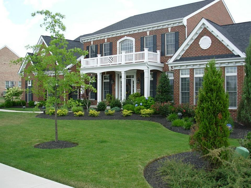Two-story brick house with a white balcony, black shutters, and well-maintained landscaping.
