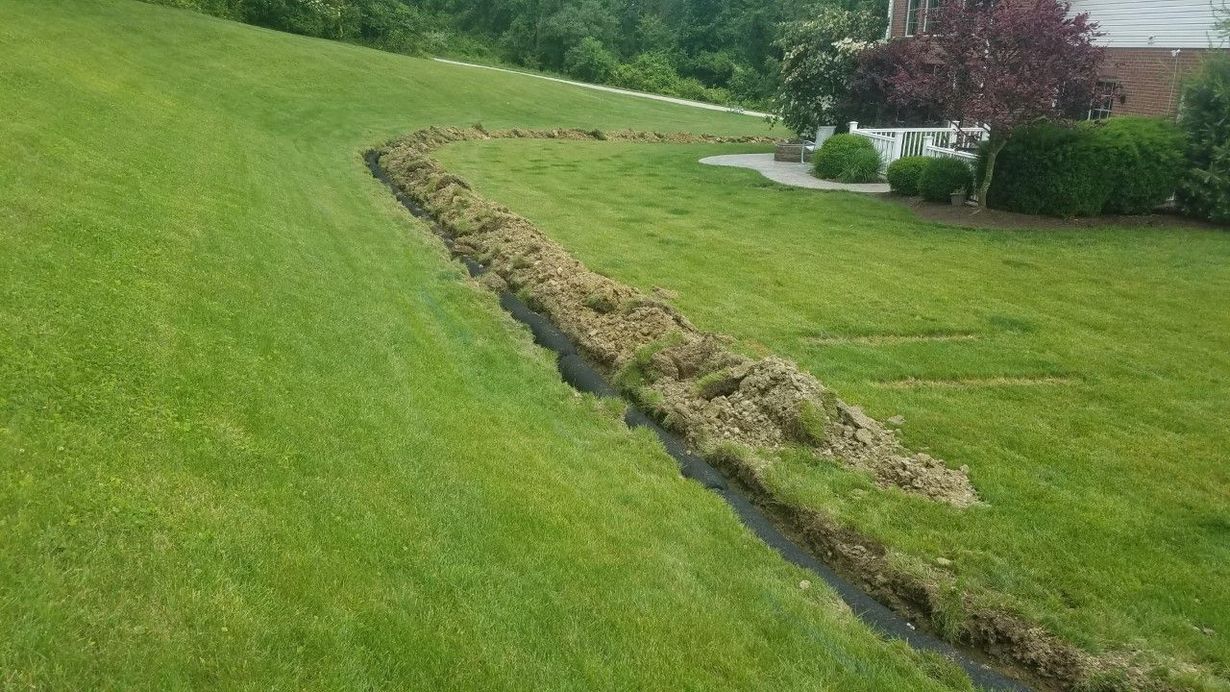 A trench dug into a green sloped lawn, with displaced earth piled along the side, leading toward a house.