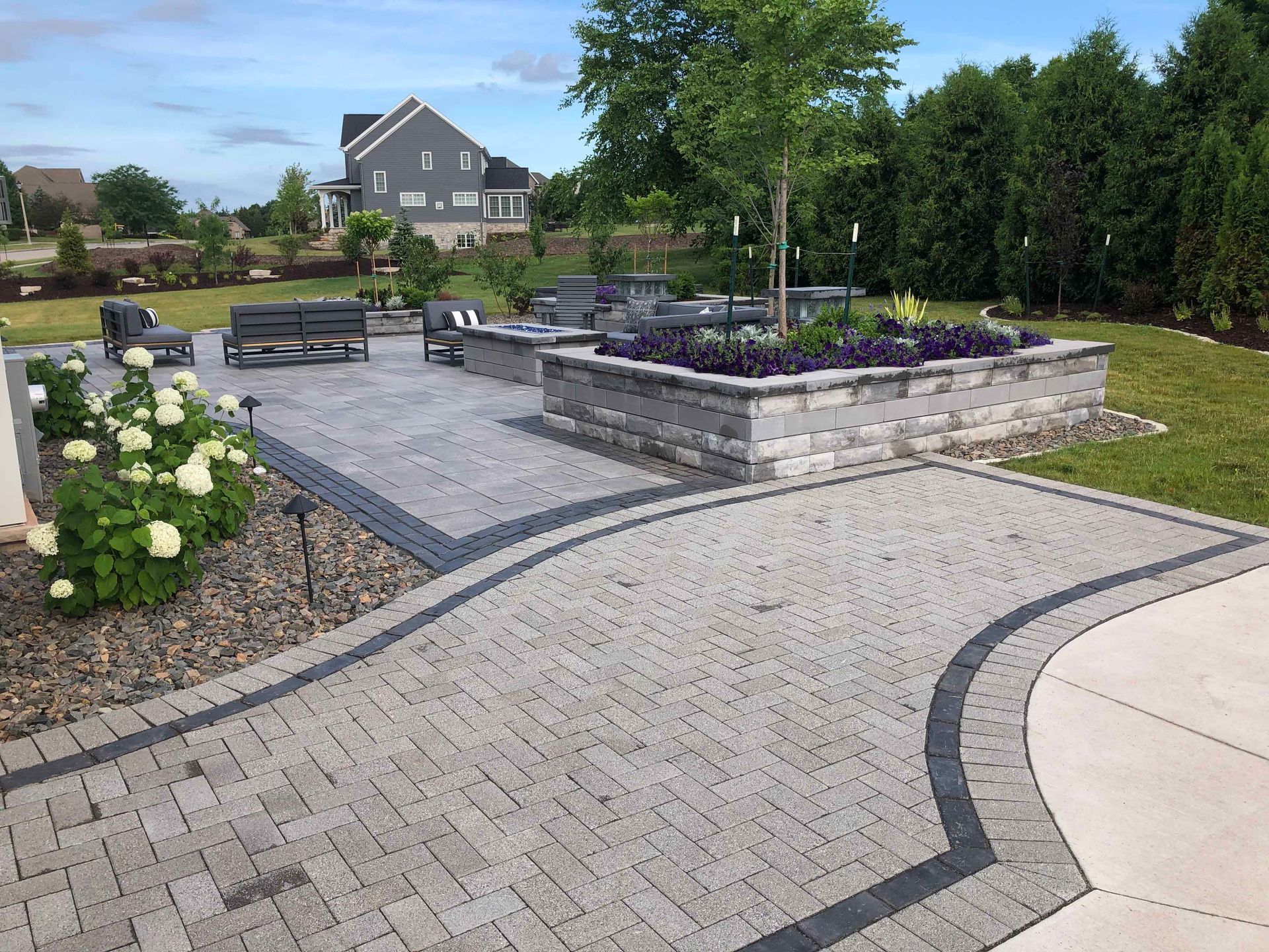 A brick walkway leading to a patio with a fire pit and flowers.