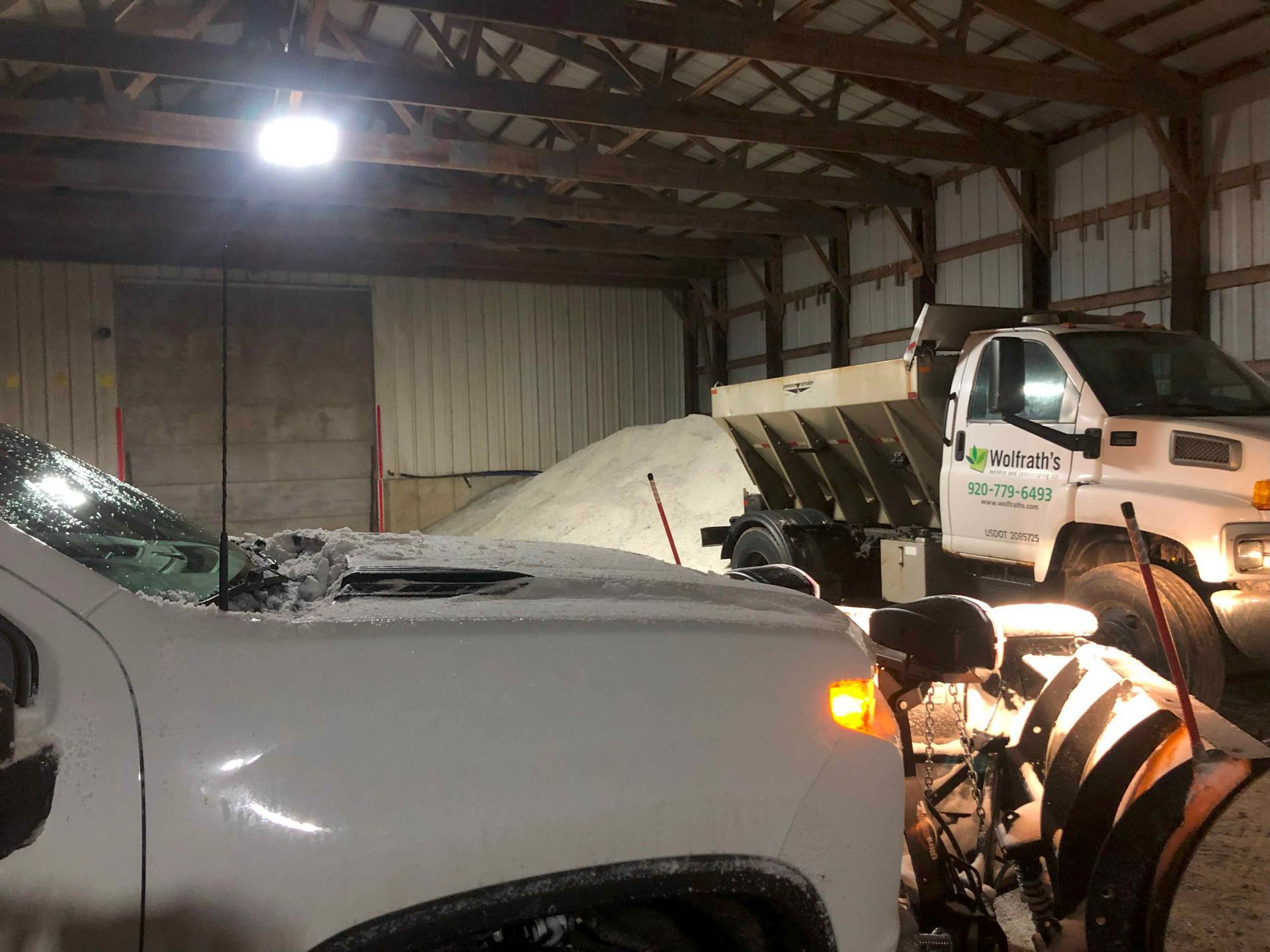 A snow plow is parked in a warehouse next to a truck.