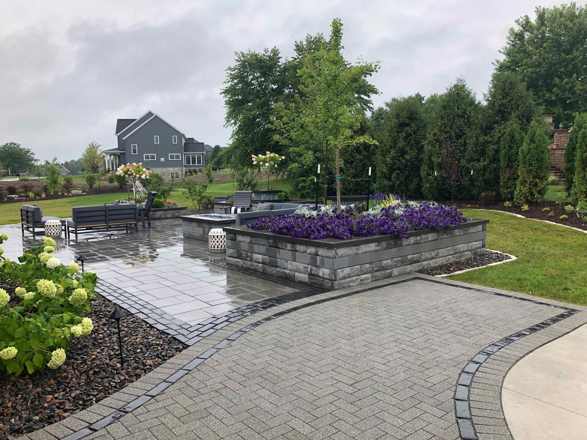 A brick walkway leading to a patio with flowers and a house in the background.