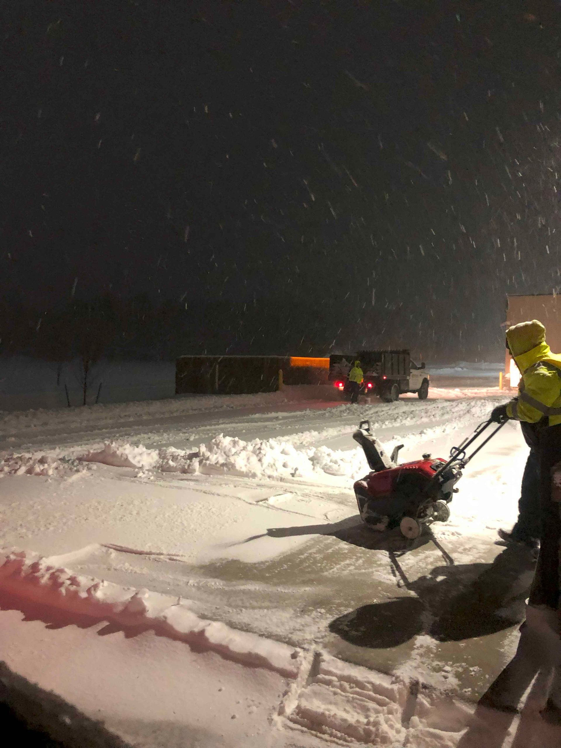A man is pushing a snow blower in the snow at night.