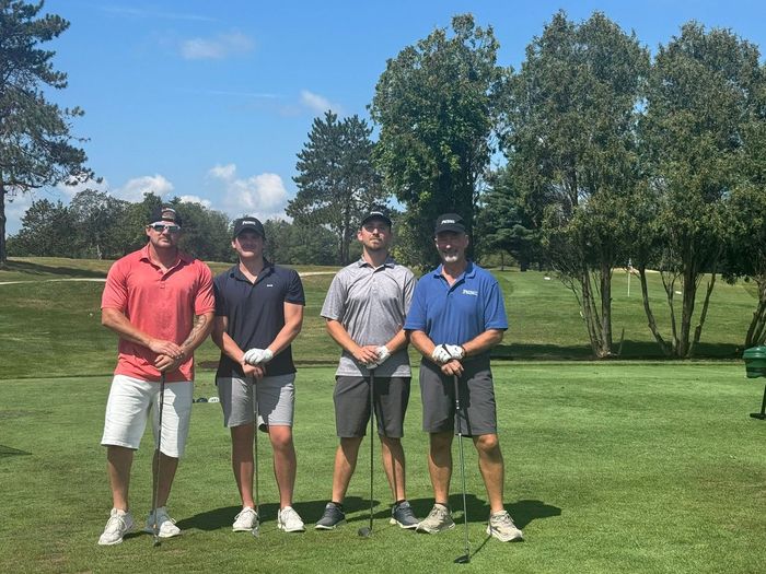 Four golfers standing on a green fairway with trees and blue sky in the background.