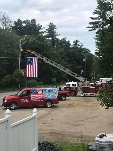 Fire trucks and a ladder truck displaying a large American flag at a roadside event outdoors.