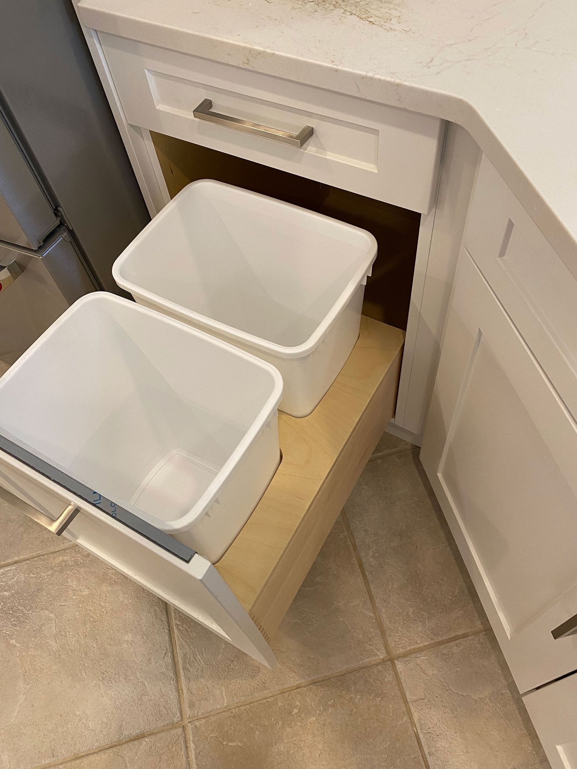 White kitchen cabinet with pull-out drawer holding two white trash bins.