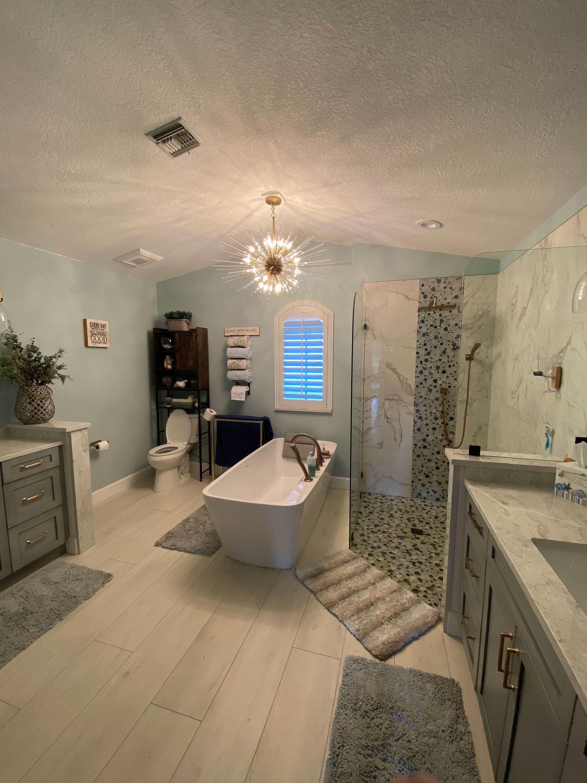 Spacious bathroom with light blue walls, white tub, shower, and vanity. A chandelier hangs from the ceiling.