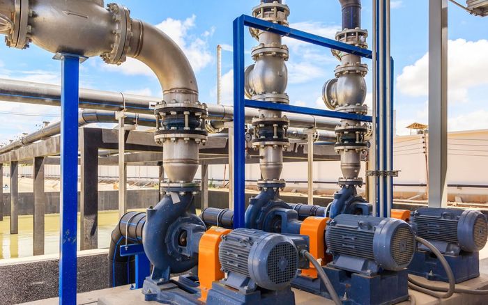 Industrial pumps with blue piping and yellow motors connected to metallic pipes at an outdoor facility under a blue sky.