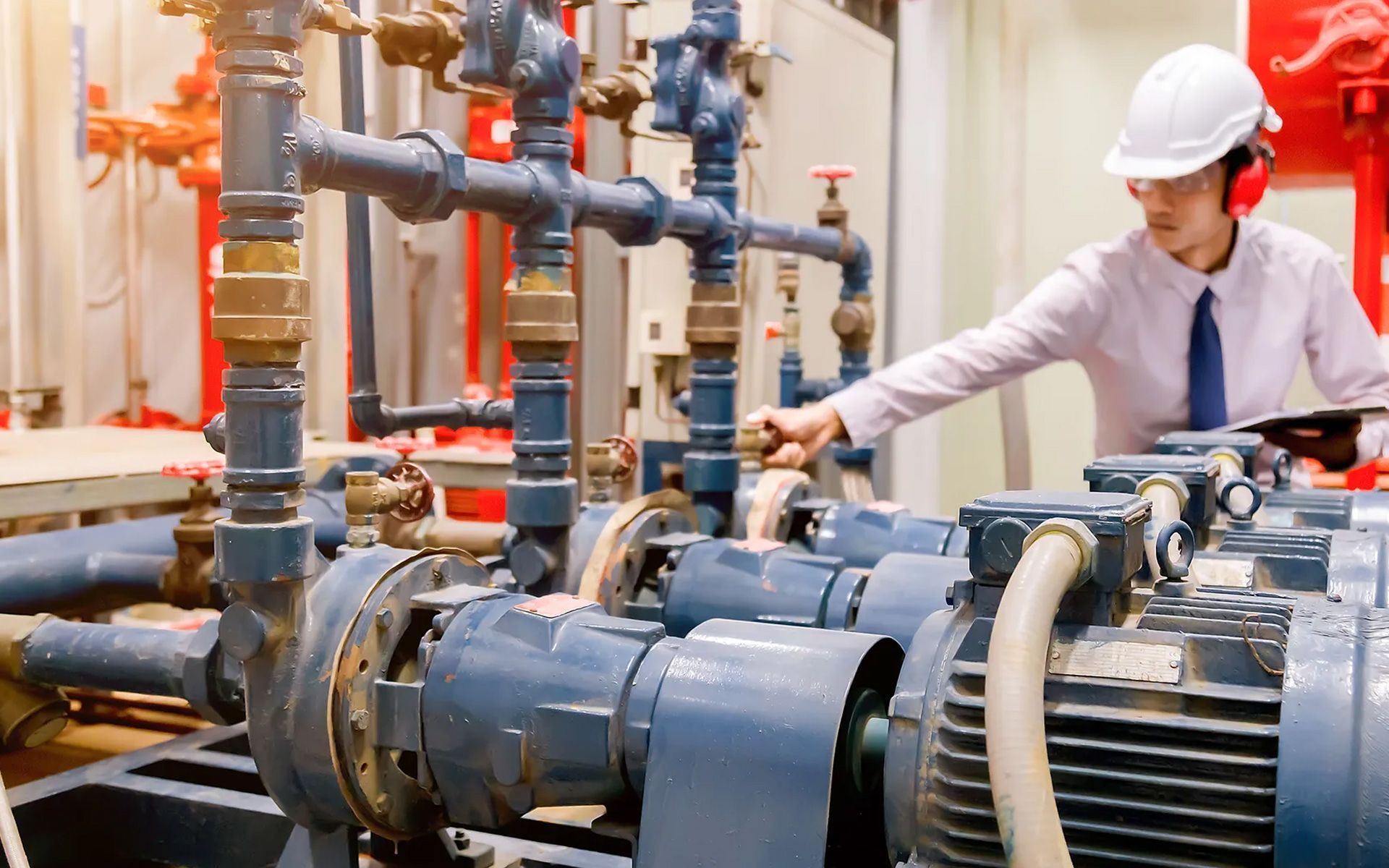 An engineer in a white hard hat and ear protection inspects industrial pipes and pumps in a manufacturing facility.