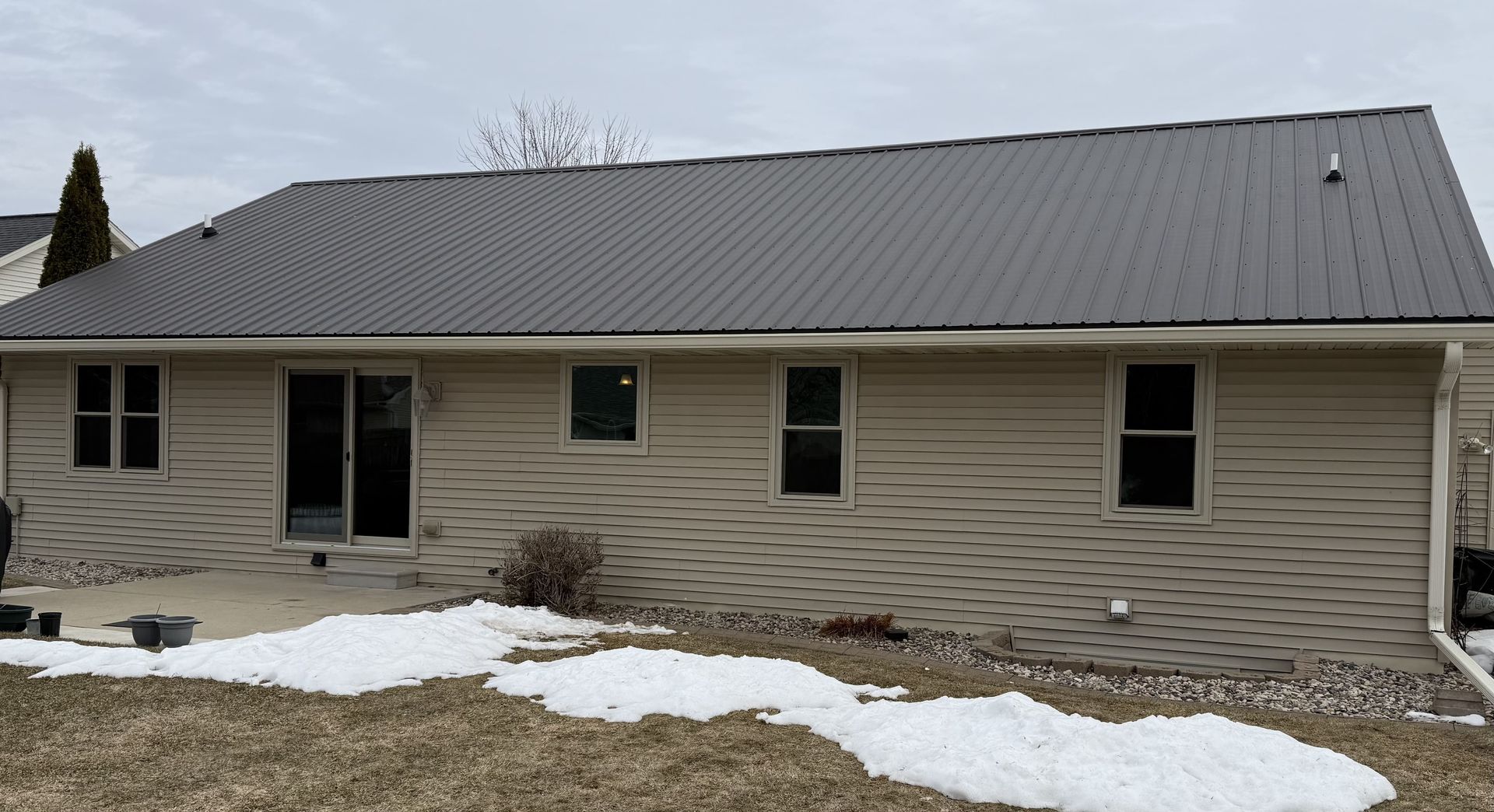 Single-story beige house with dark gray roof and snow patches in the yard