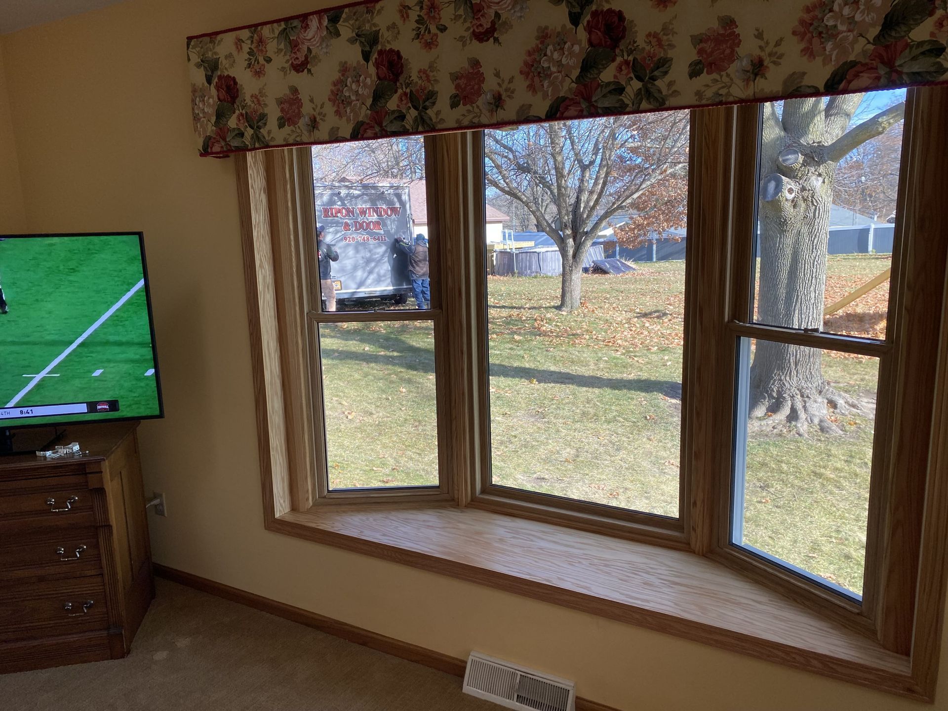 Living room bay window with view of yard, TV on left, and floral valance above curtains